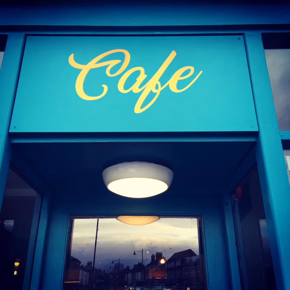 The cafe sign over the door at 87 Market Place at dusk, gold script on deep teal, the square reflected in the glass