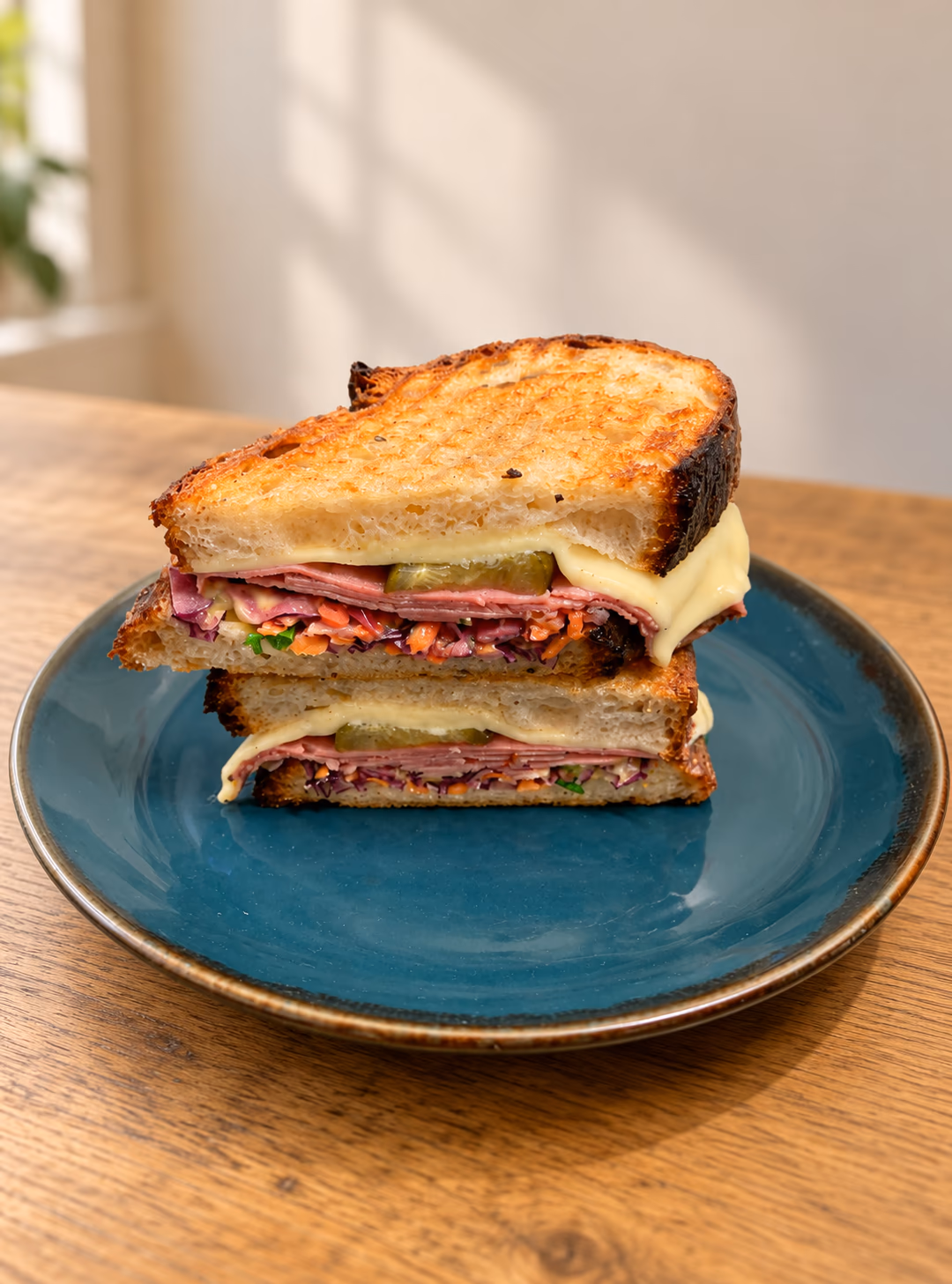 Two toasted Reuben halves stacked on a dark teal plate, showing melted cheese, pastrami, slaw and gherkin