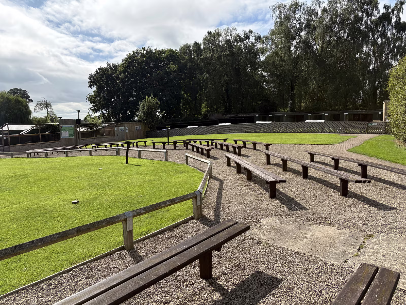 Rows of wooden benches on gravel curving round the grass flying ground, with the aviaries under the trees behind