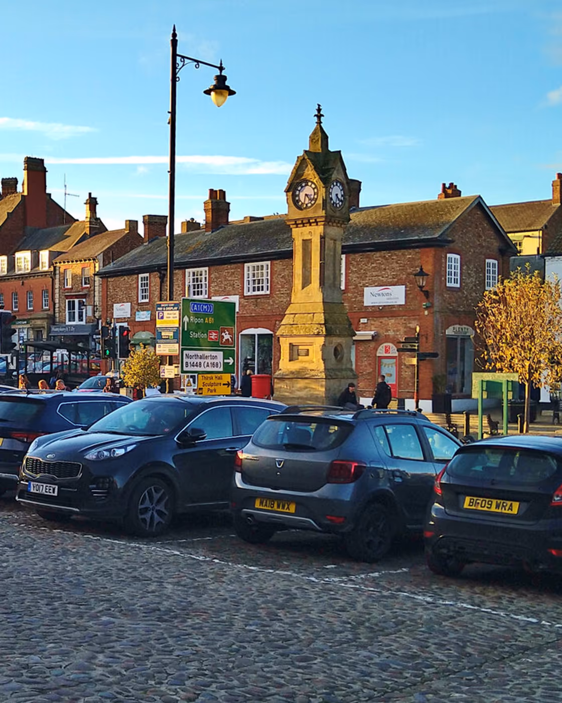 The Victorian clock memorial in Thirsk market square, with period buildings, a lamp post and parked cars on the cobbles