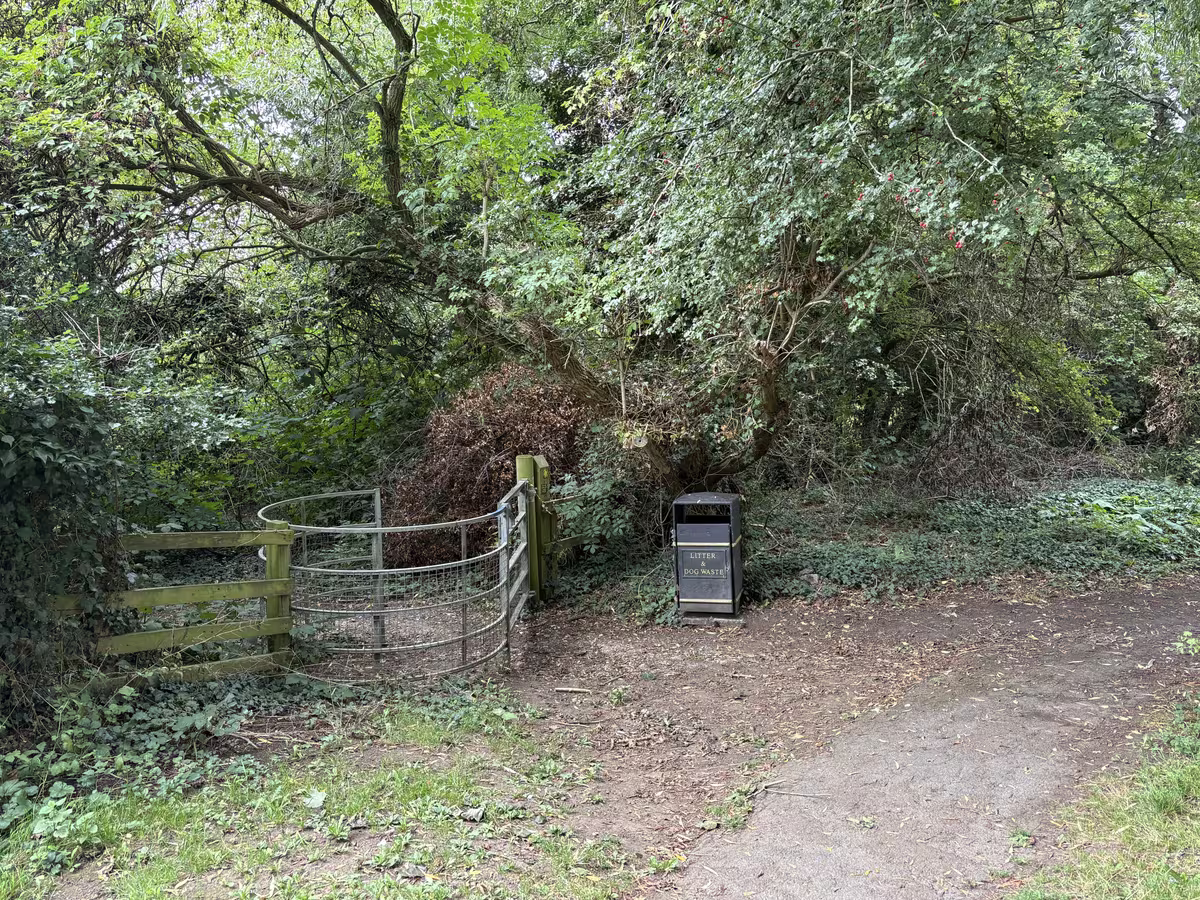 The metal kissing gate at the Stockton Road end, a litter bin beside it and the path running out through the trees