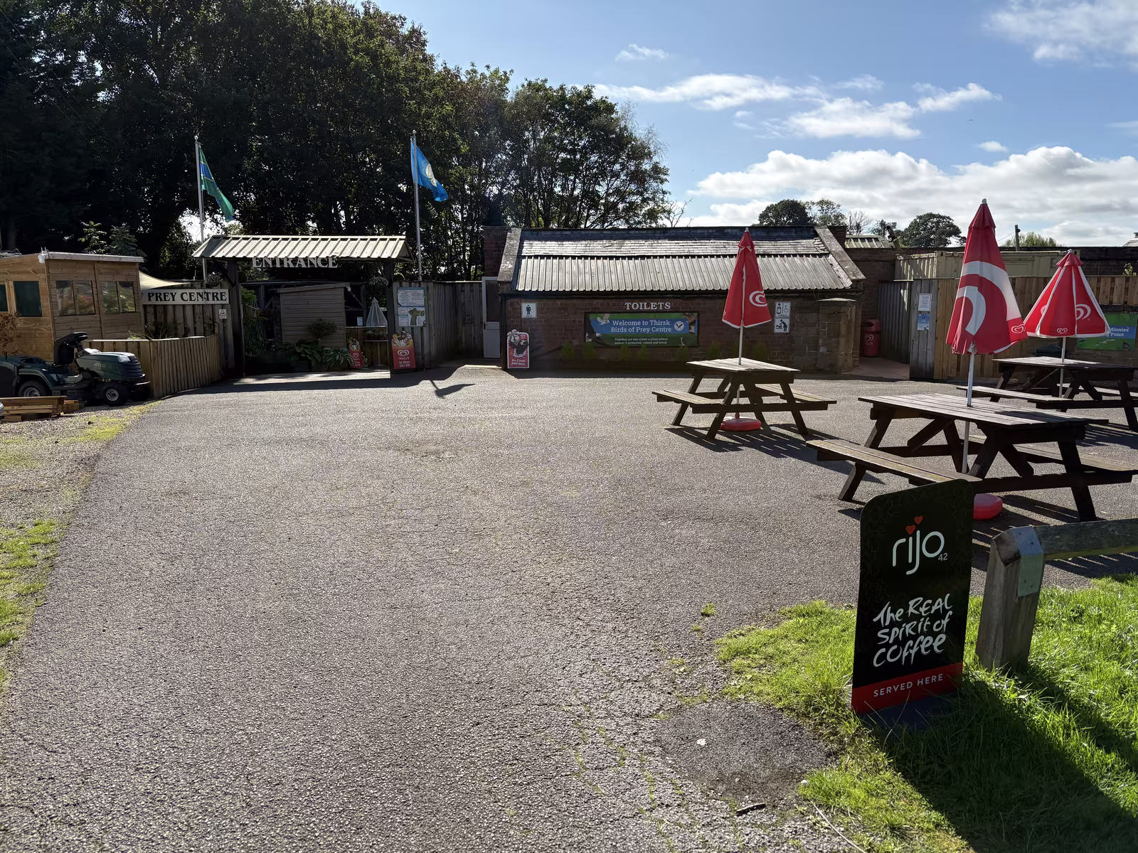 The way in: the entrance on the left, the toilets straight ahead, and picnic tables under red umbrellas on the tarmac beside a sign for coffee